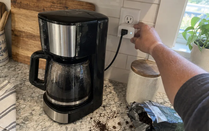 Hand plugging a smart plug with a coffee maker attached into a kitchen outlet